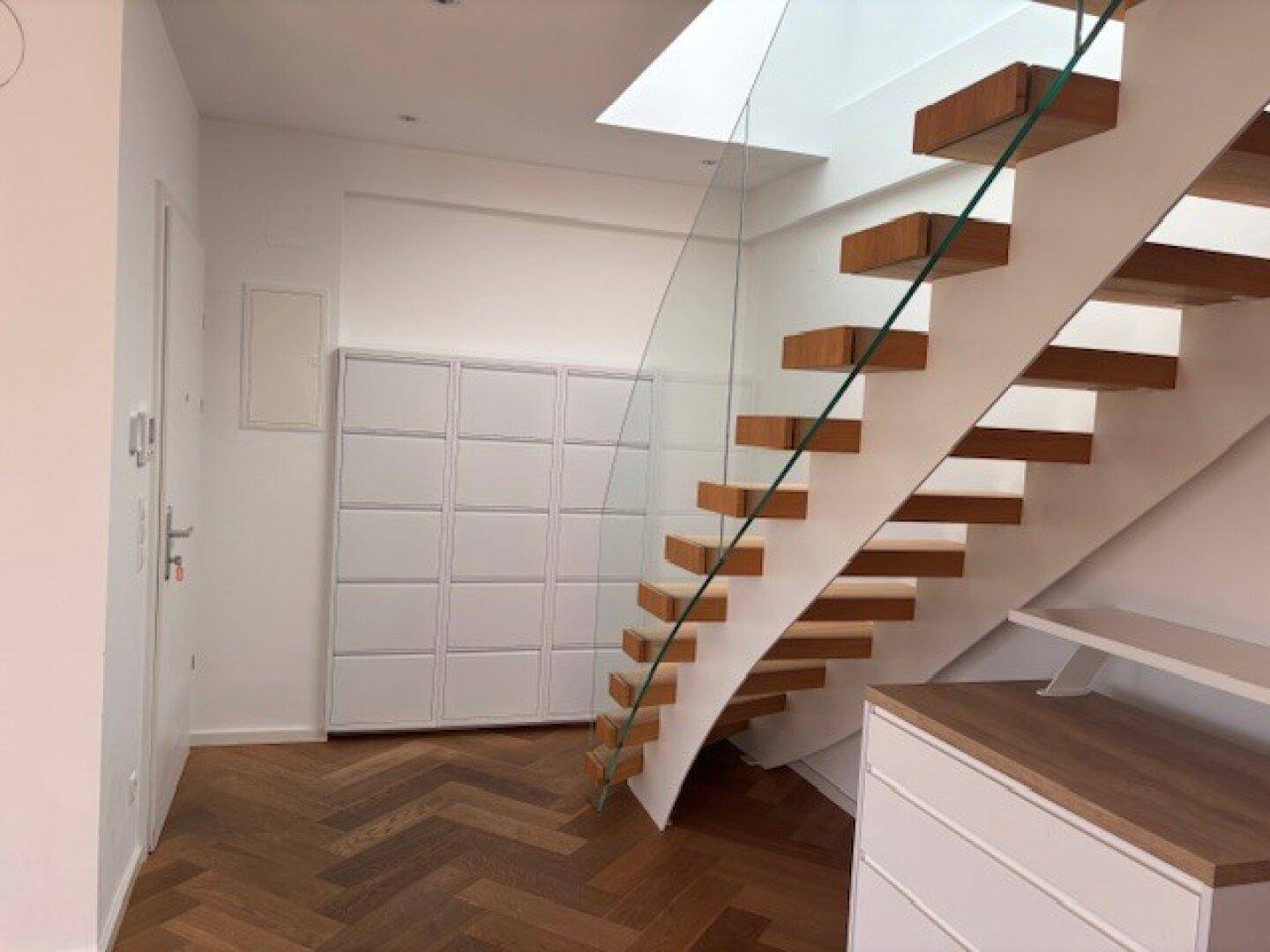 Eingangsbereich mit moderner Holztreppe, Glasgeländer und Fischgrätparkettboden.