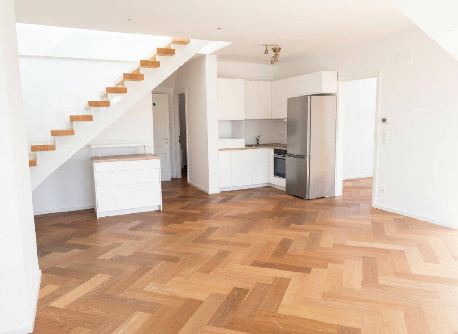 Offener Wohnbereich mit moderner Küchenzeile, Holztreppe und Fischgrätparkettboden.