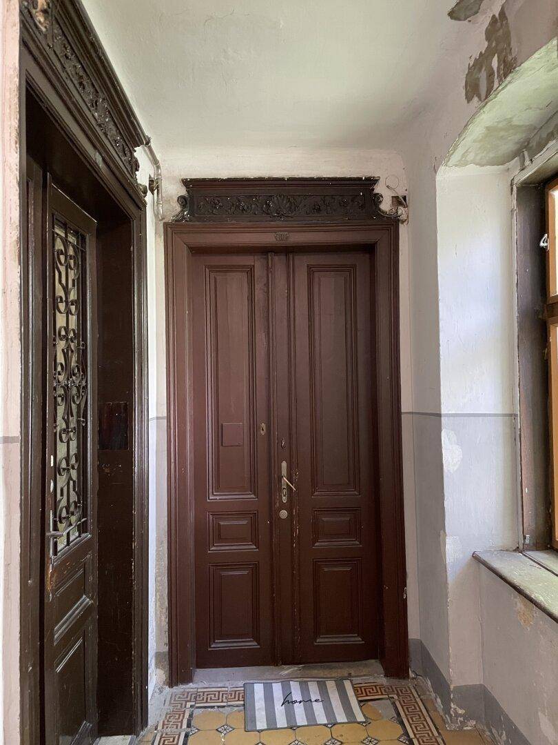 Eingangstür aus dunklem Holz in einem Flur mit historischen Fliesen und Tageslicht.