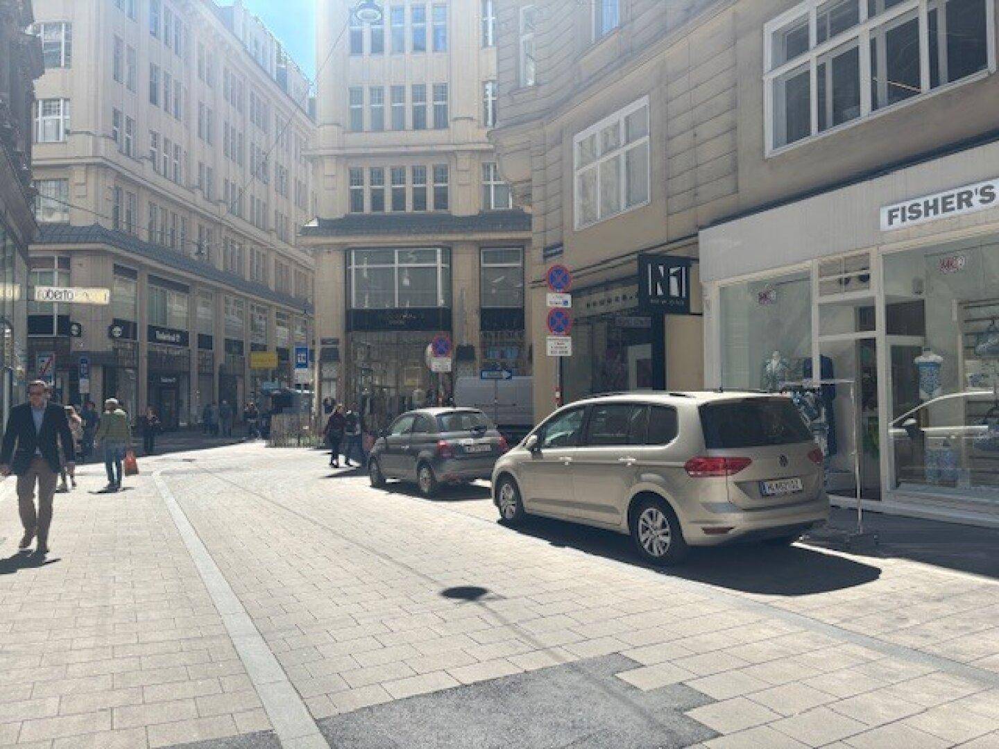 Sonnige Stadtstraße mit geparkten Autos und Passanten, umgeben von klassischen Geschäftsgebäuden in Wien.