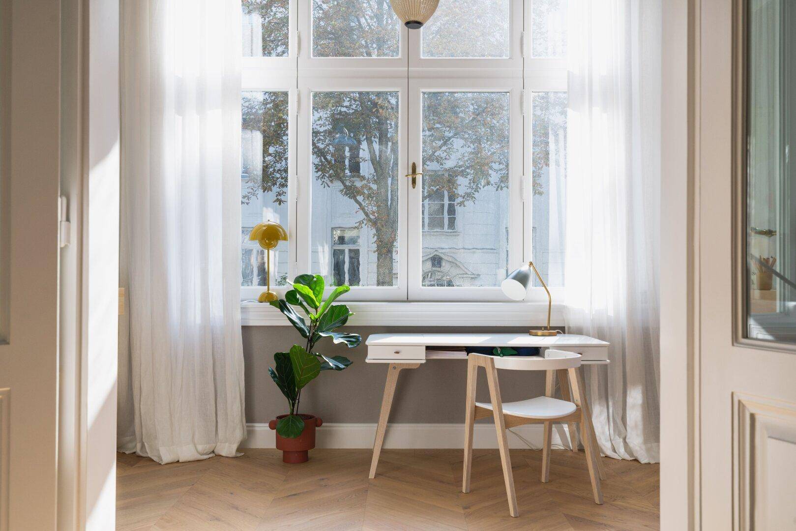 Helles Arbeitszimmer mit Schreibtisch am Fenster, Holzboden und viel Tageslicht.