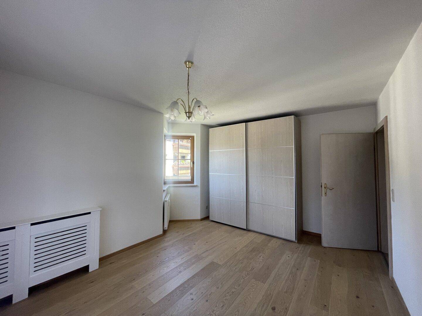 Schlafzimmer mit Holzboden, Fenster und einem großen Schrank, gut beleuchtet.