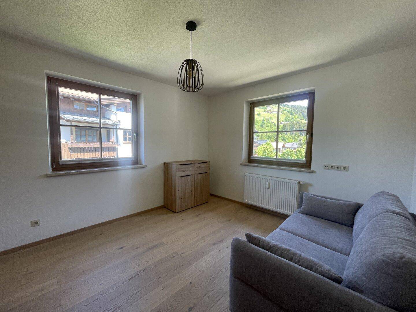 Gemütliches Zimmer mit Holzboden, Sofa und zwei Fenstern mit Blick ins Grüne.