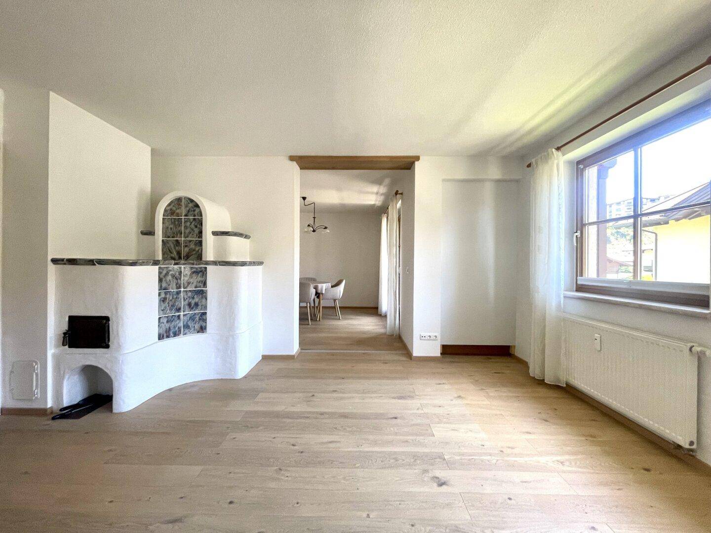 Helles Wohnzimmer mit hellem Holzboden und traditionellem Kachelofen, offen zum Essbereich.