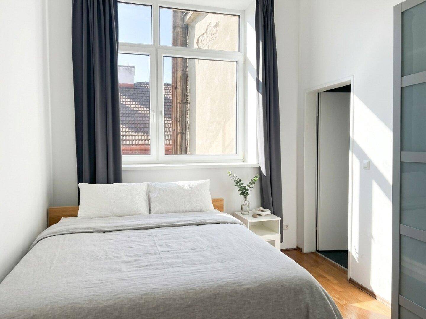 Helles Schlafzimmer mit Holzboden, großem Fenster und dunklen Vorhängen.