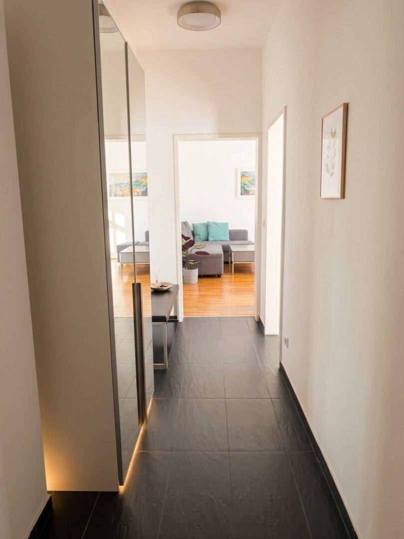 Heller Flur mit dunklen Fliesen, Spiegelschrank und Blick ins Wohnzimmer.