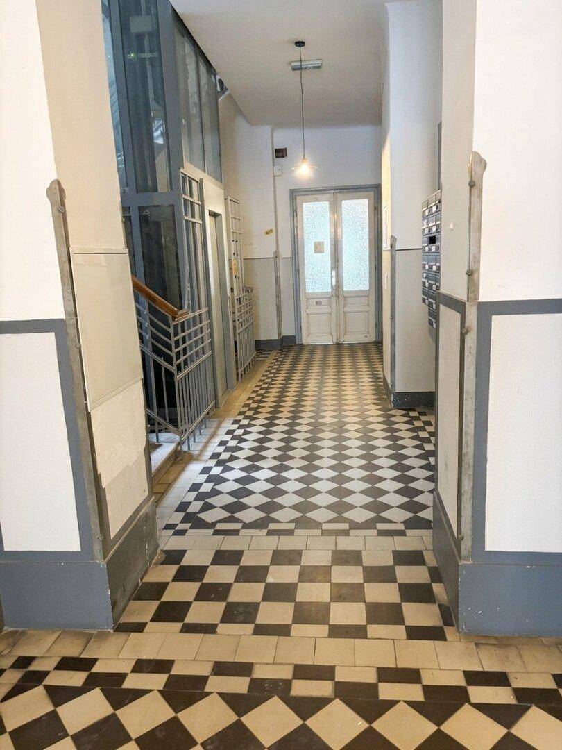 Langer Flur in einem Altbau mit Schachbrettmuster-Fliesen und Aufzug.
