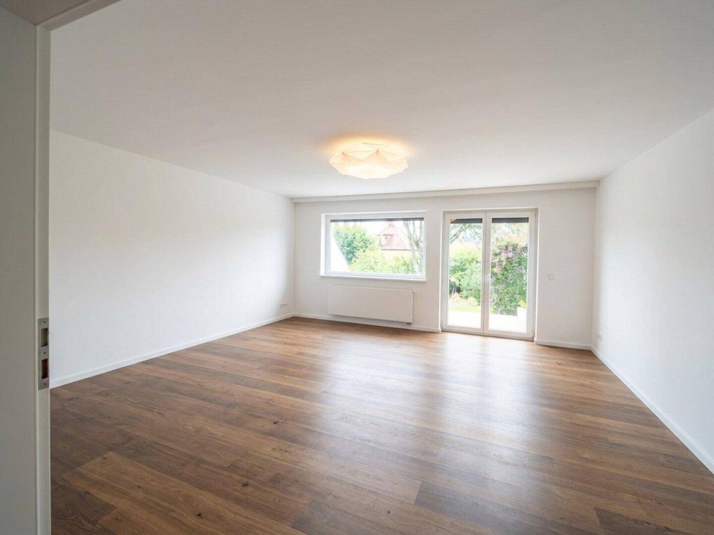 Leerer, heller Raum mit Holzboden und großen Fenstern zum Garten.