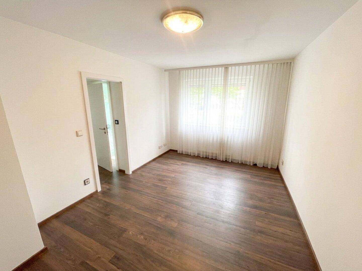 Heller, leerer Raum mit dunklem Holzboden und Fenster mit Vorhängen.