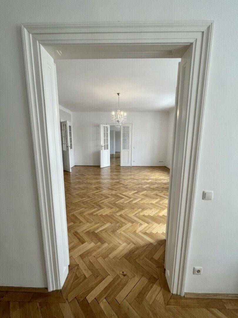Elegante Flügeltüren führen in einen hellen Raum mit Fischgrätparkett und Kronleuchter.