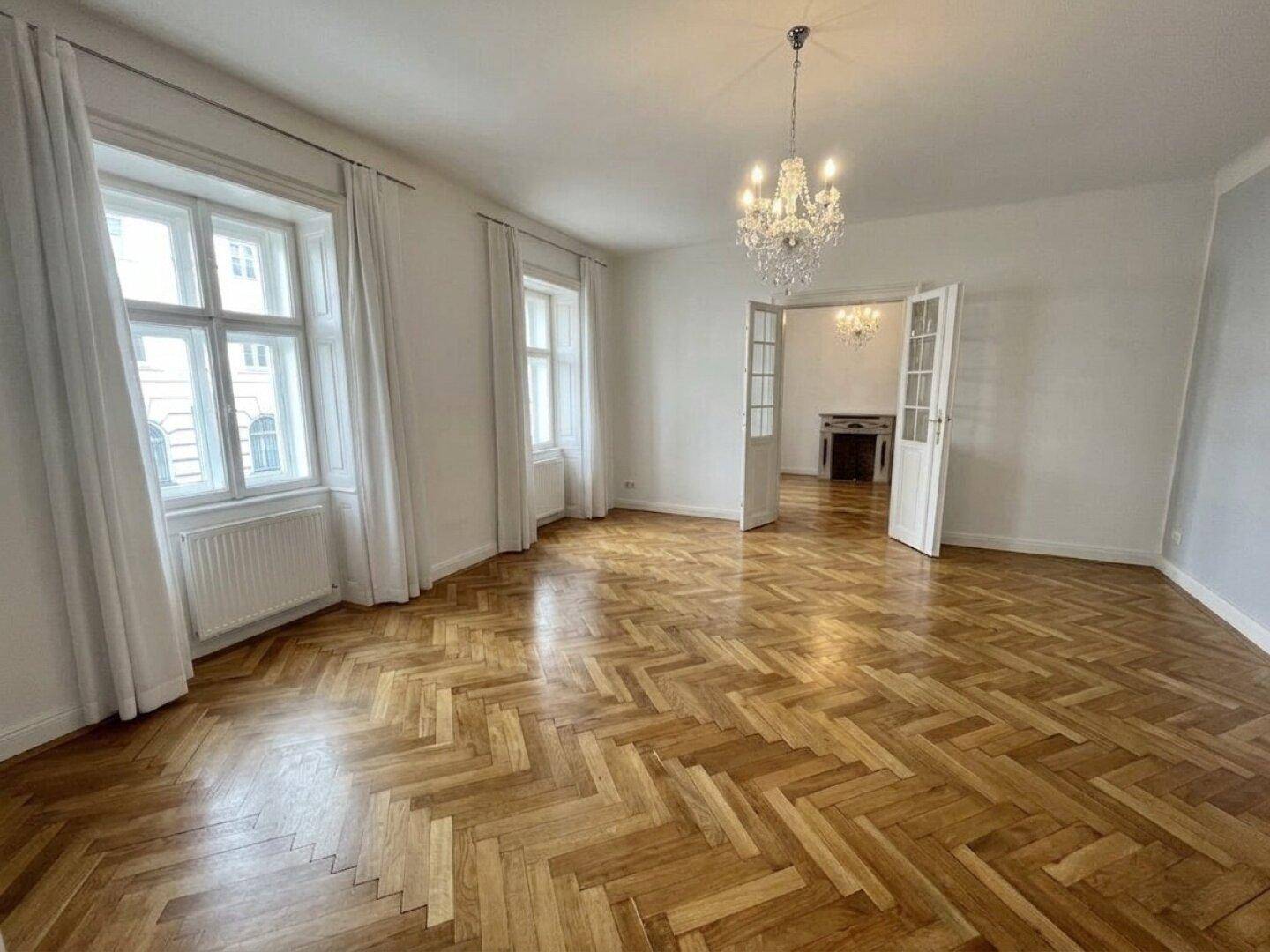 Helles Wohnzimmer mit klassischem Fischgrätparkett, hohen Fenstern und einem dekorativen Kamin.