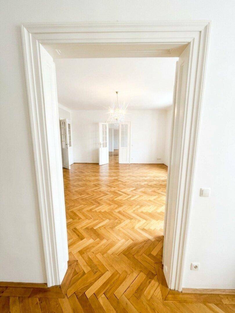 Blick durch eine breite, verzierte Türöffnung in einen hellen Raum mit klassischem Fischgrätparkettboden.