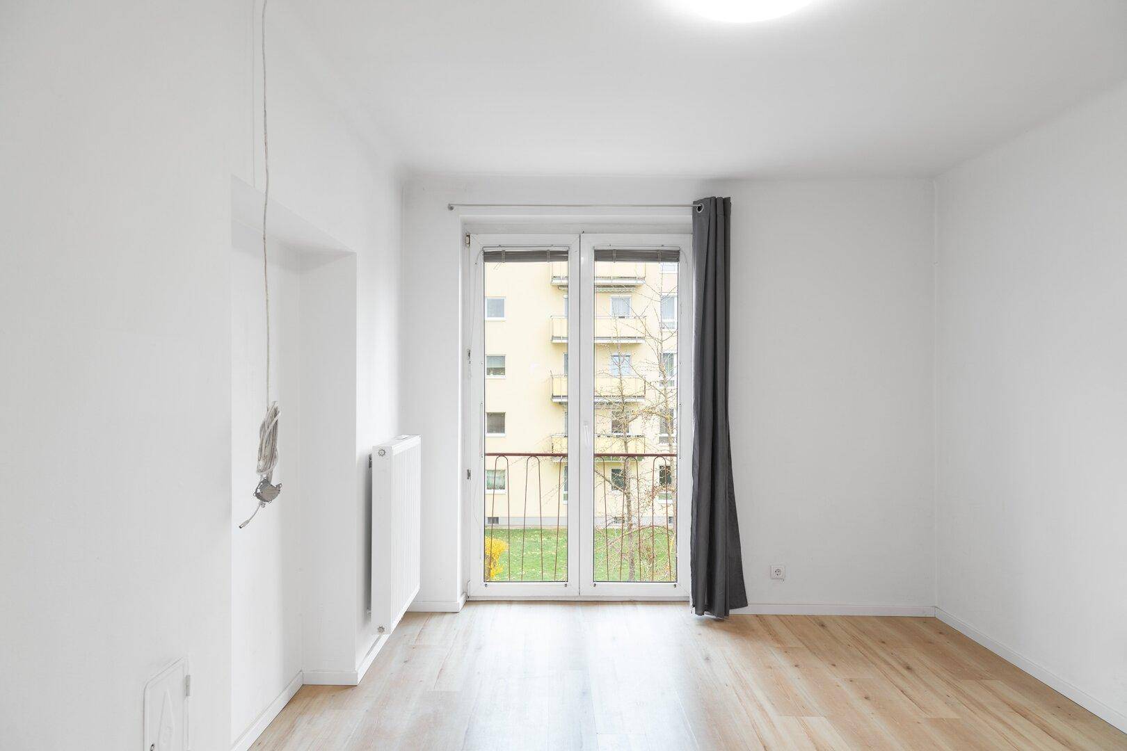 Lichtdurchfluteter Raum mit hellem Holzboden und Zugang zu einem kleinen Balkon mit Blick auf den Innenhof.