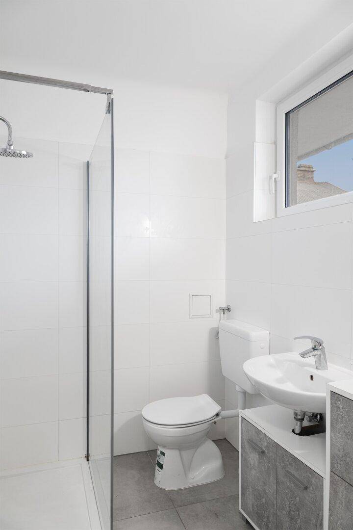 Modernes Badezimmer mit weiß gefliesten Wänden, Dusche, WC, Waschbecken und einem Fenster für Tageslicht.