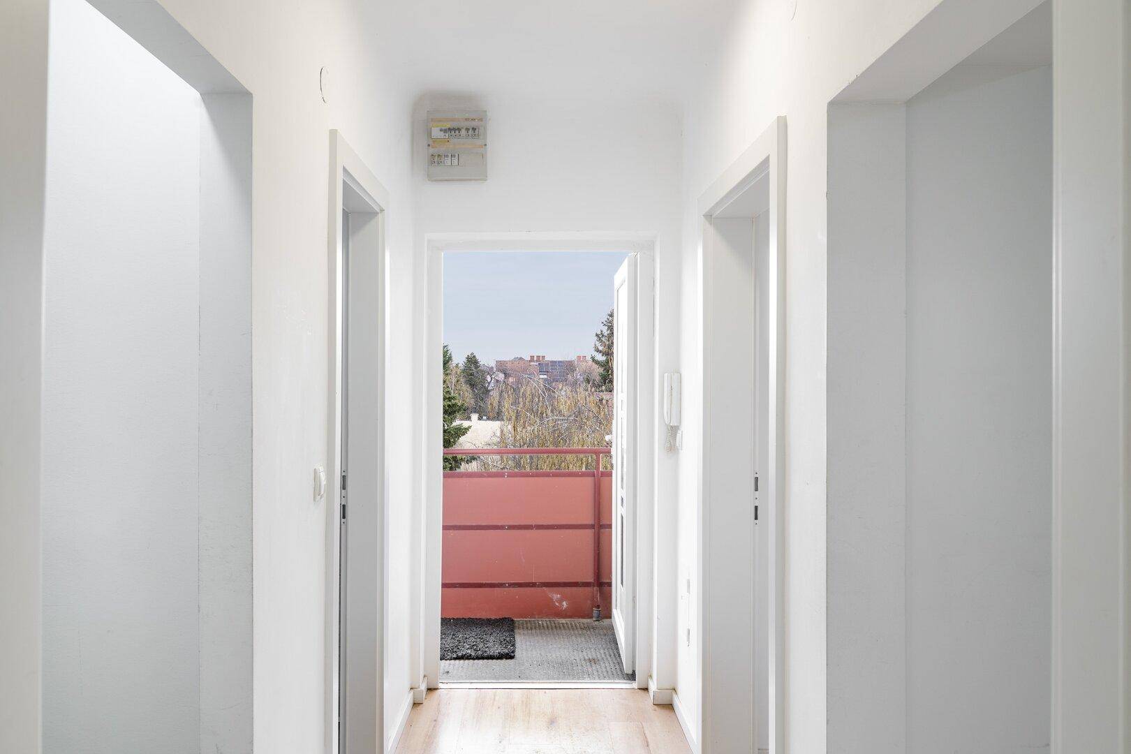 Heller Flur mit Holzboden, der zu einem Balkon mit rotem Geländer und Fernblick führt.