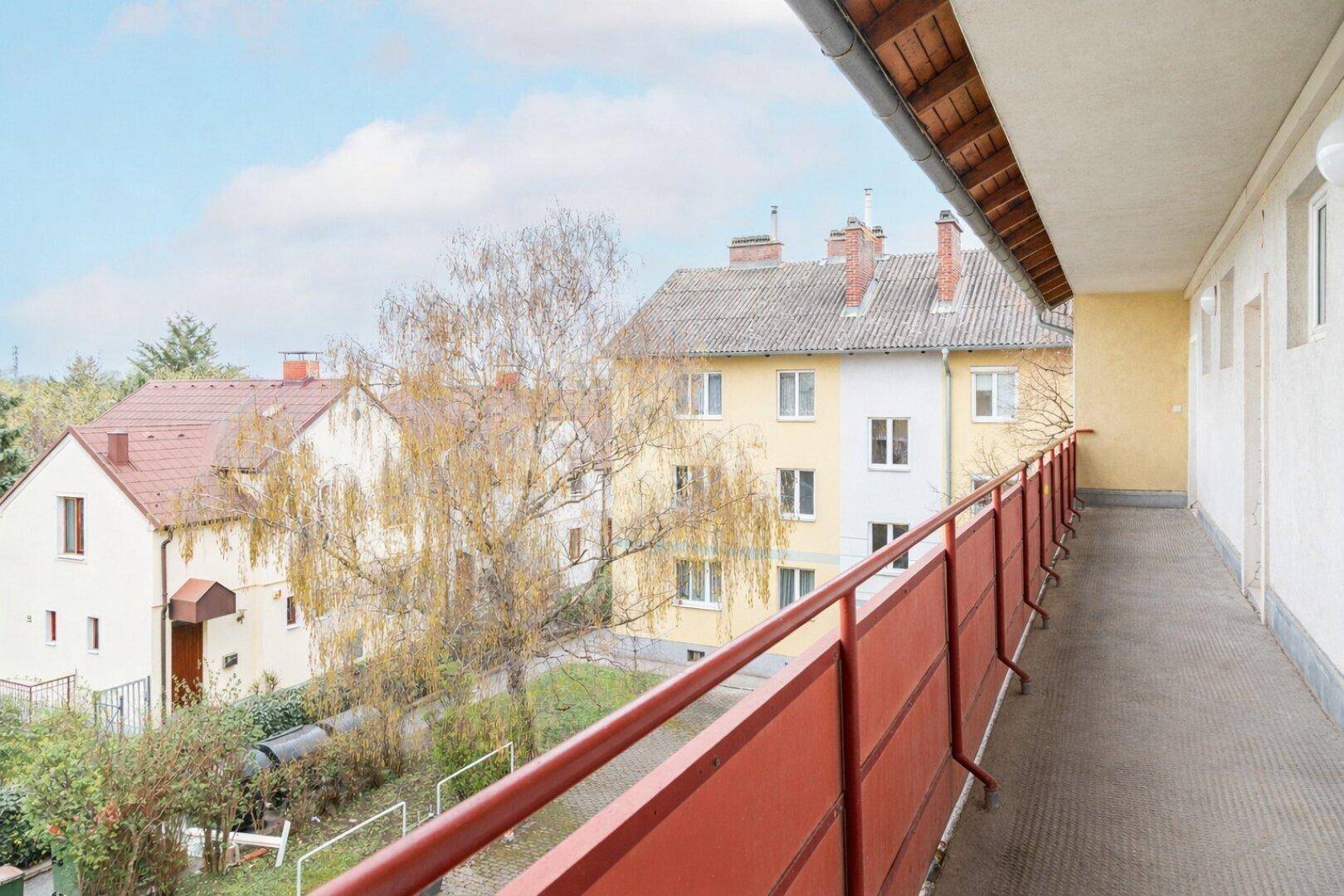 Langer Balkon mit rotem Geländer und Blick auf den grünen Innenhof und die umliegenden Wohnhäuser.