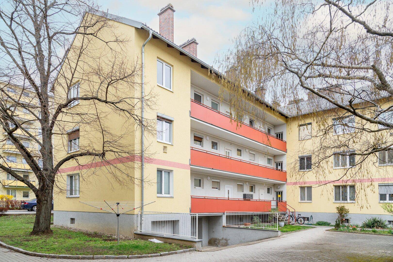 Gelbes Mehrfamilienhaus mit roten Balkonen, umgeben von Bäumen und einer kleinen Grünfläche.
