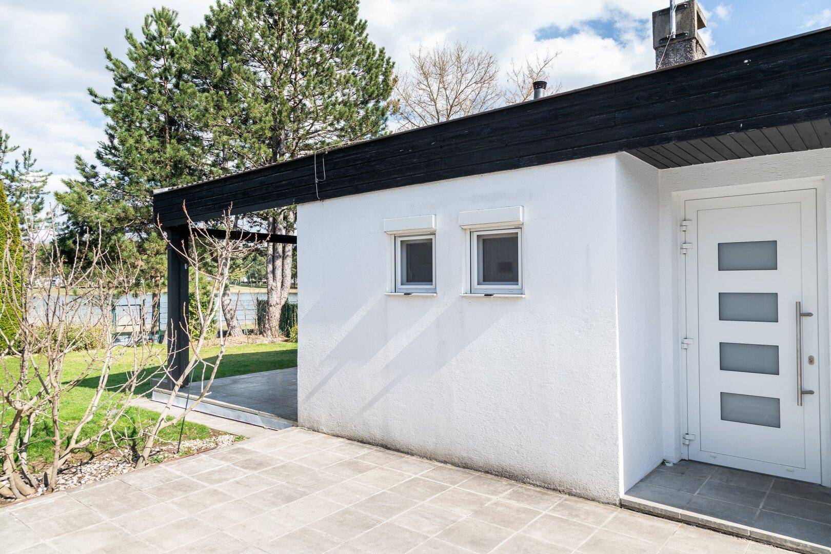 Eingangsbereich des Hauses mit weißer Fassade, moderner Tür und gefliester, überdachter Terrasse.