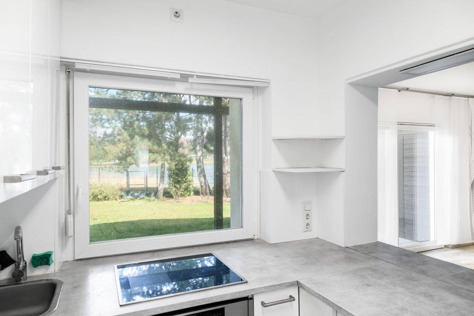 Moderne Küche mit Induktionskochfeld, weißen Schränken und Blick auf den Garten und See.