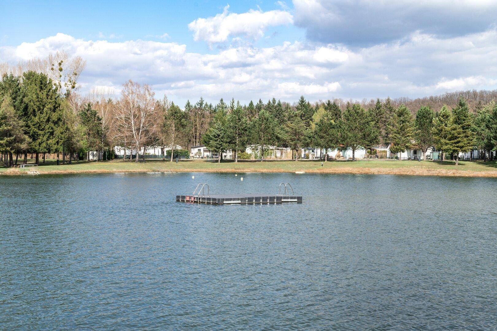 Panoramablick auf einen ruhigen See mit schwimmender Plattform und Häusern am gegenüberliegenden Ufer.
