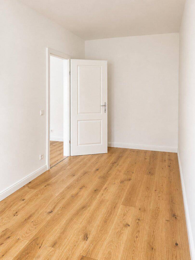 Leerer Raum mit Holzboden und weißer Tür, die zu einem weiteren Zimmer führt.