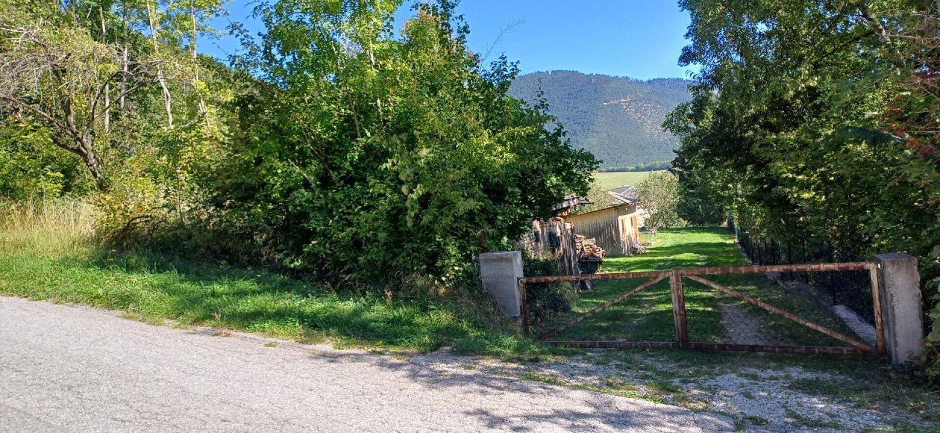 Einfahrt zum Grundstück mit Holztor, umgeben von üppigem Grün und einem weiten Blick auf die Berge.