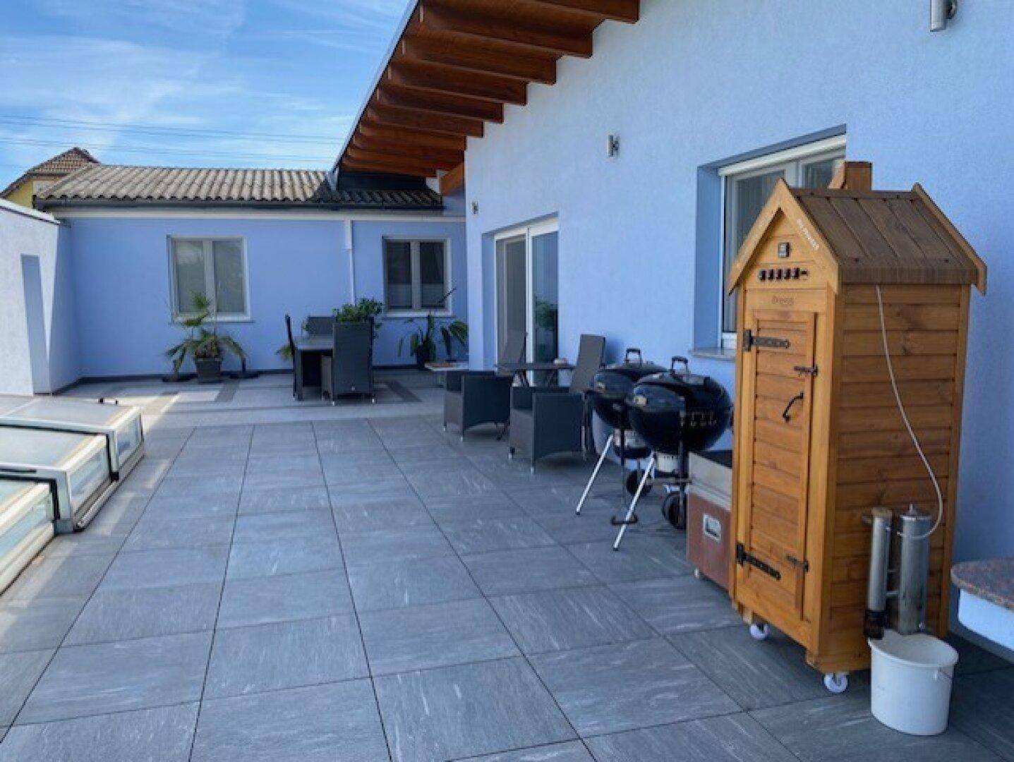 Große geflieste Terrasse mit Grillbereich und Holzräucherofen vor dem Haus.