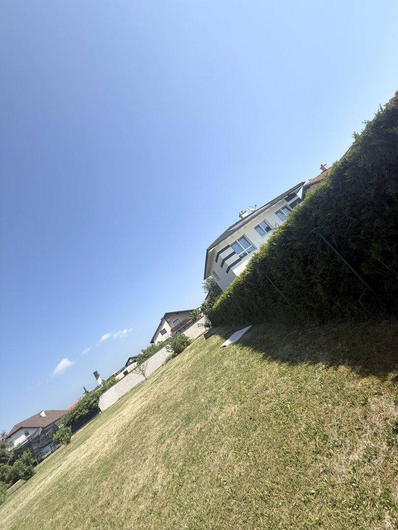 Abfallendes Gartengrundstück mit gepflegter Hecke und Blick auf ein Wohnhaus unter blauem Himmel.