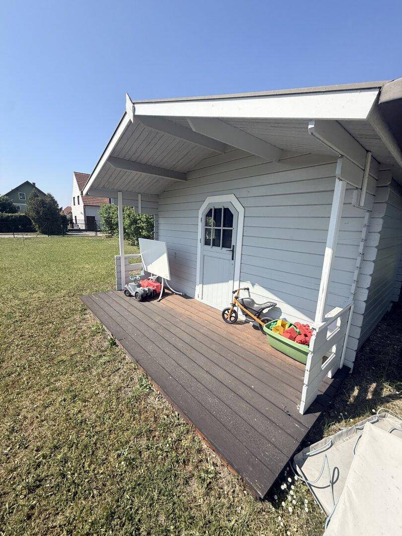 Kleines weißes Gartenhaus mit Veranda und Kinderspielzeug auf einer grünen Rasenfläche.