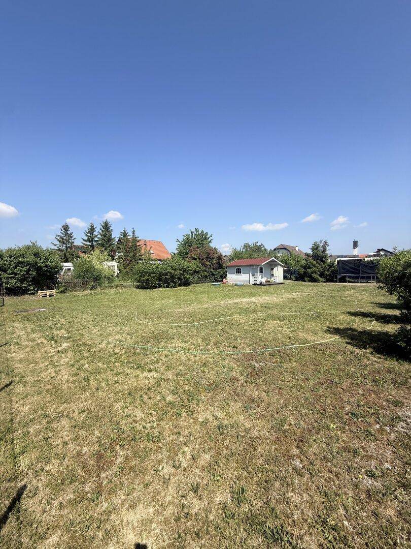 Weitläufiges Gartengrundstück mit Rasenfläche und einem kleinen Gartenhaus im Hintergrund unter blauem Himmel.