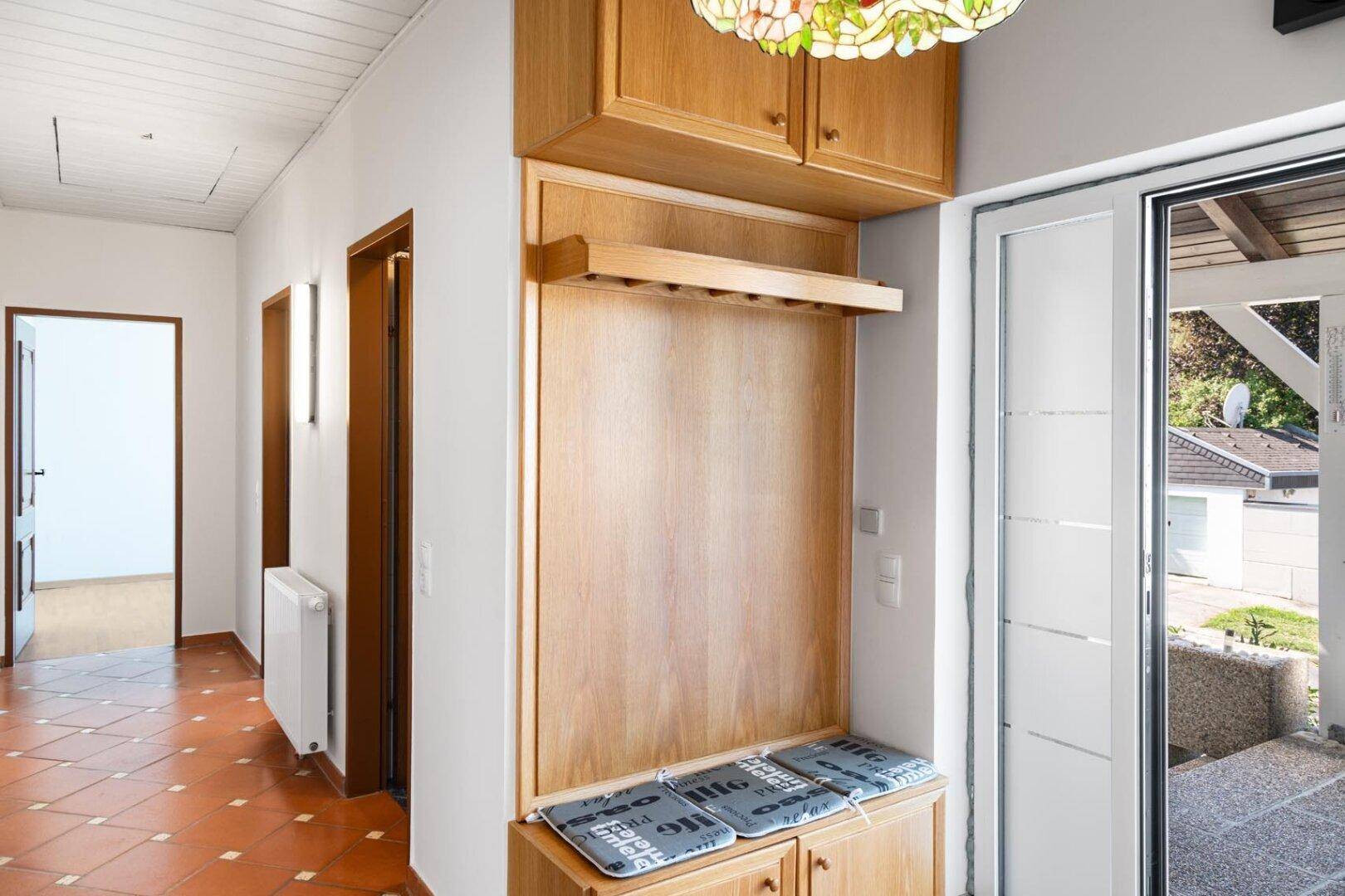 Heller Eingangsbereich mit Holzgarderobe, Terrakottafliesen und Blick nach draußen.