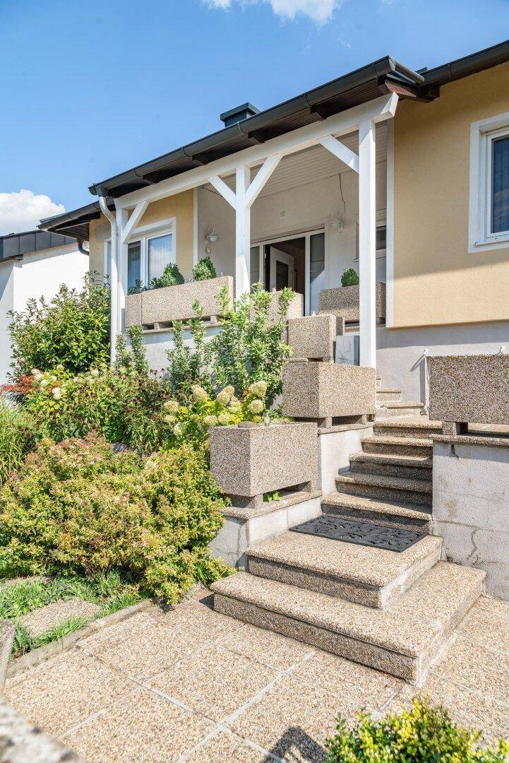 Überdachter Eingangsbereich mit Treppe, Pflanzenkübeln und sonnigem Ambiente.