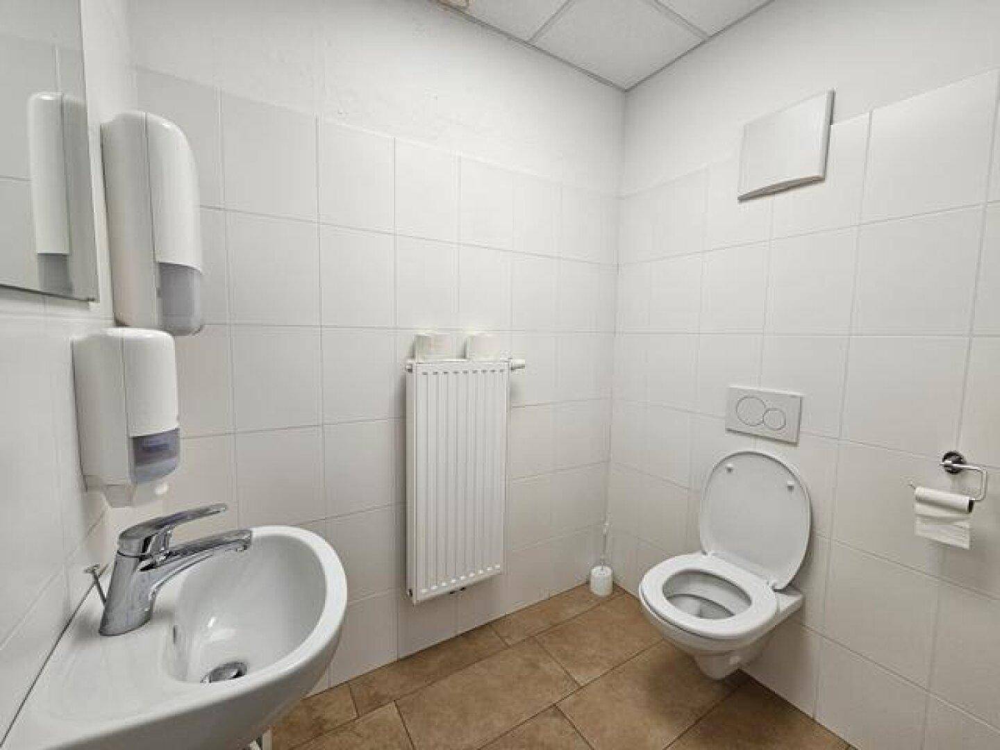 Helle Toilette mit Waschbecken, WC und weißen Wandfliesen, ausgestattet mit Seifenspendern.