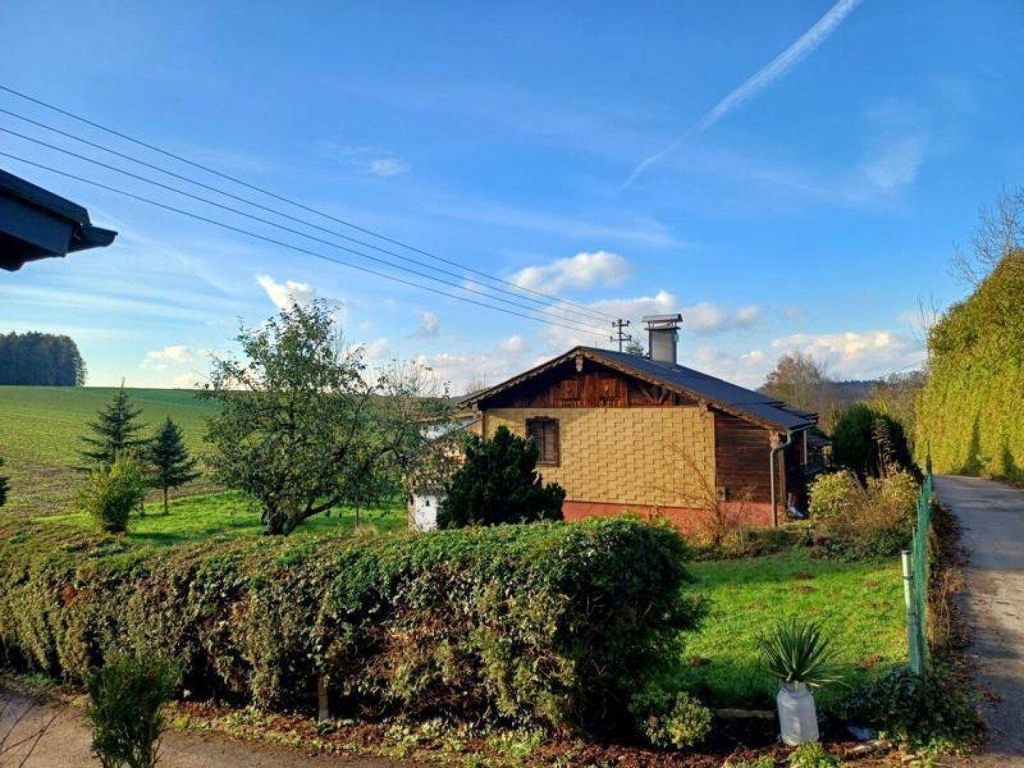 Hausfassade mit einer gepflegten Hecke im Vordergrund und weiten Feldern in der Ferne.