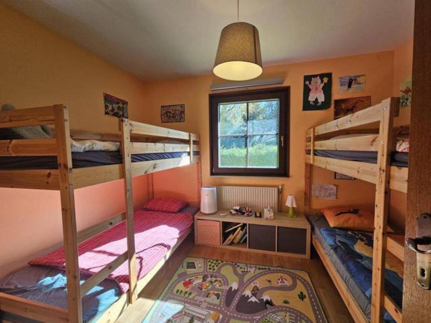 Kinderzimmer mit zwei Etagenbetten, Holzboden und farbenfrohem Teppich unter dem Fenster.