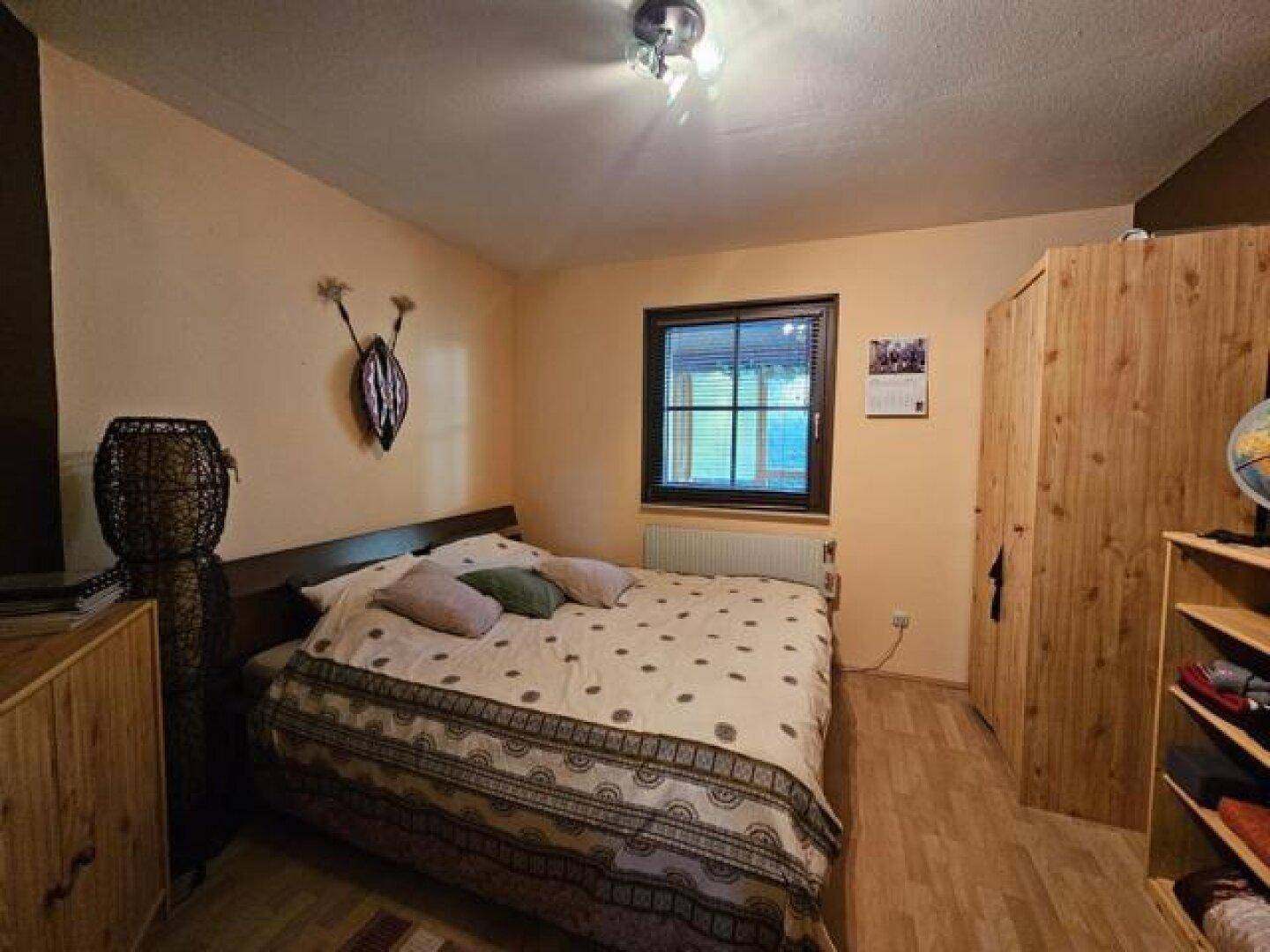Schlafzimmer mit Doppelbett, Holzschrank und Laminatboden unter einem Fenster.