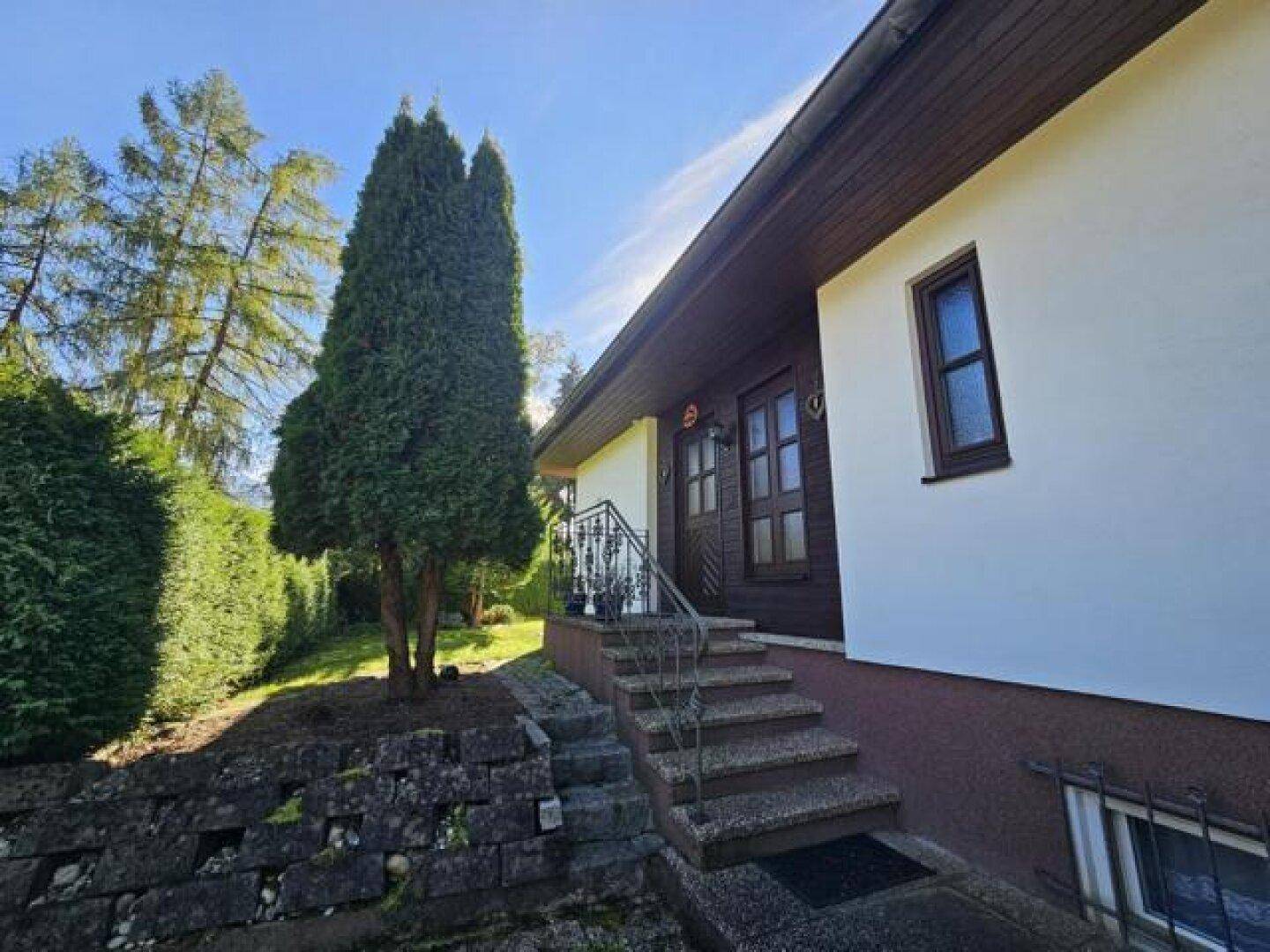 Eingangsbereich des Hauses mit Steintreppe, Geländer und umgebender Bepflanzung.