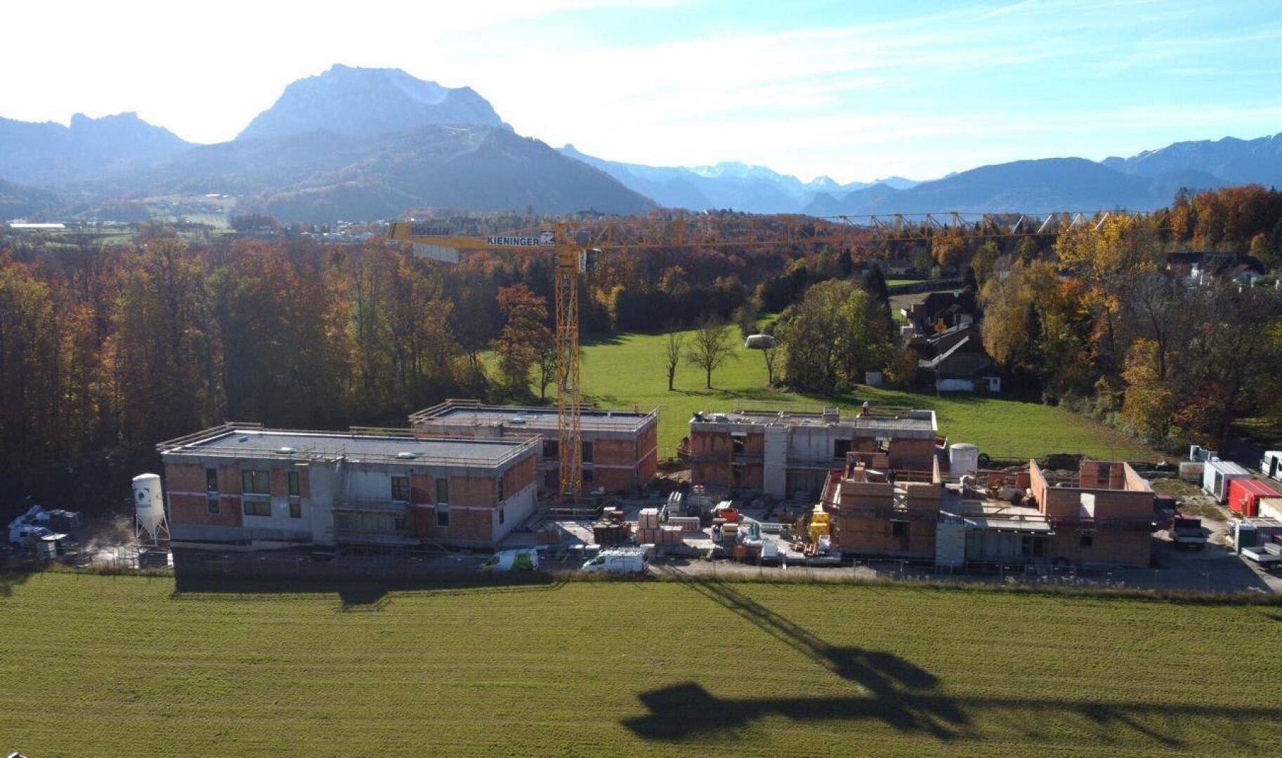 Luftaufnahme der Baustelle mit im Bau befindlichen Gebäuden, umgeben von Bäumen, Bergen und einem See in der Ferne.