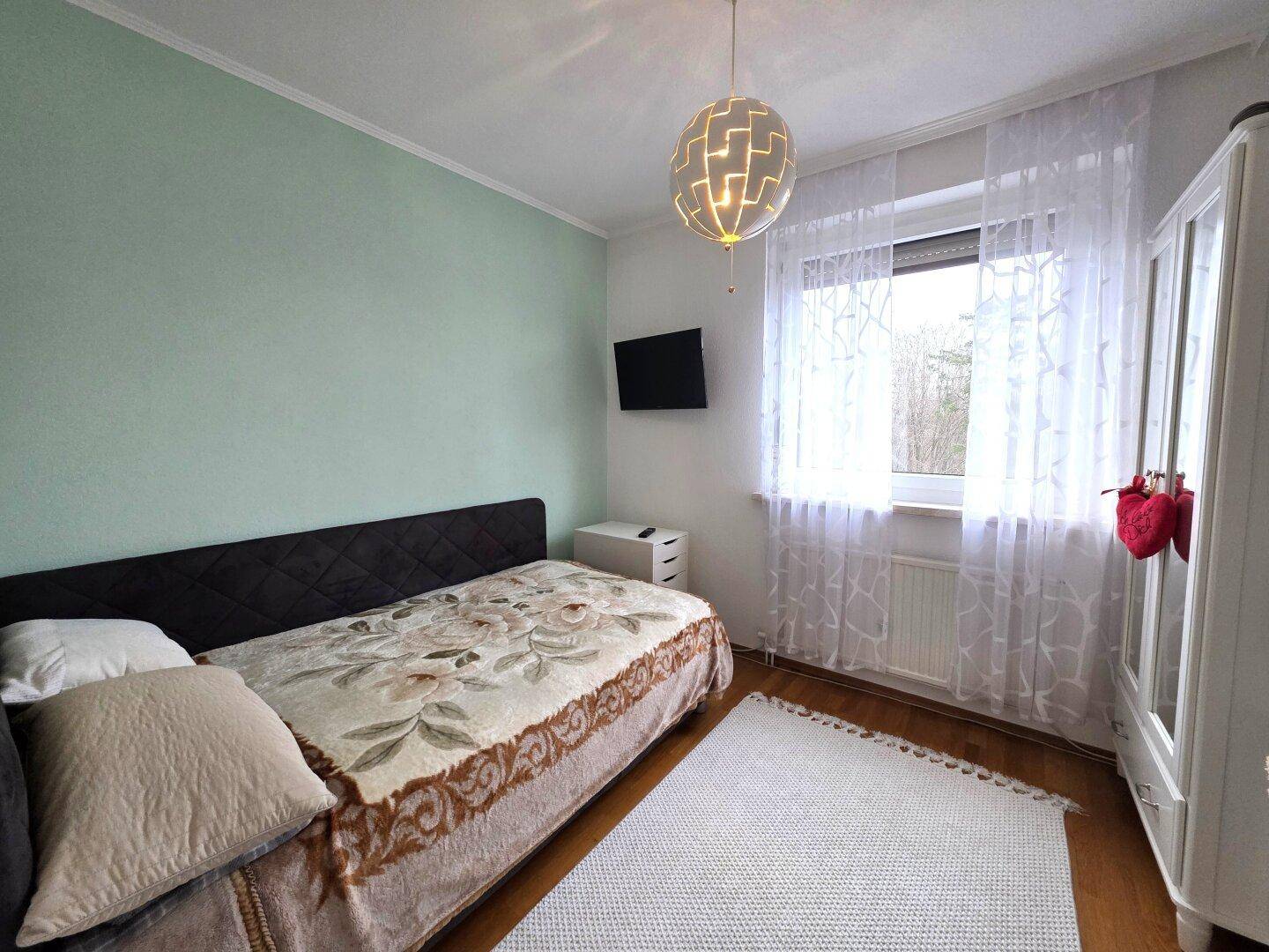 Helles Schlafzimmer mit Einzelbett, Fernseher und Fenster mit Vorhängen.