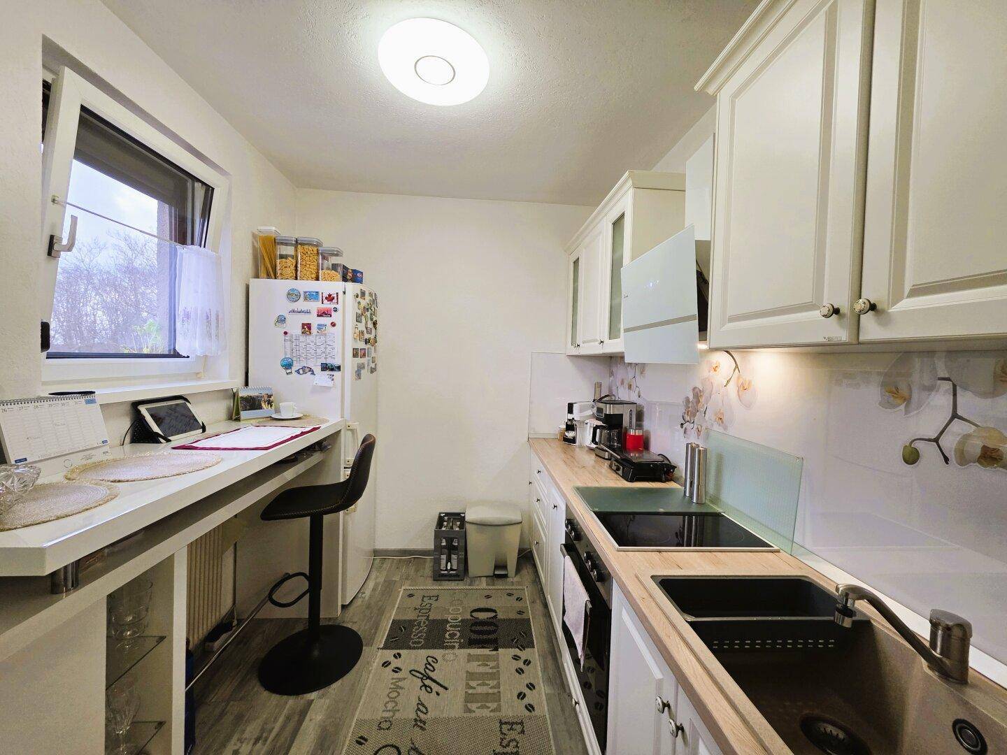 Funktionale Küche mit weißen Schränken, Kühlschrank und Fenster mit Blick ins Grüne.