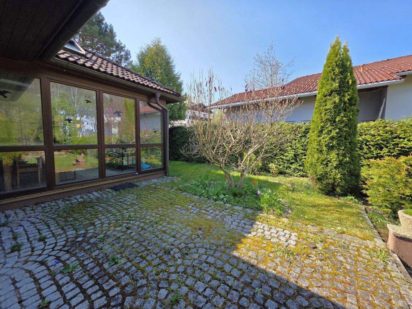 Gepflasterte Terrasse neben einem Wintergarten, umgeben von Grünflächen und einer Hecke, ideal für sonnige Stunden.