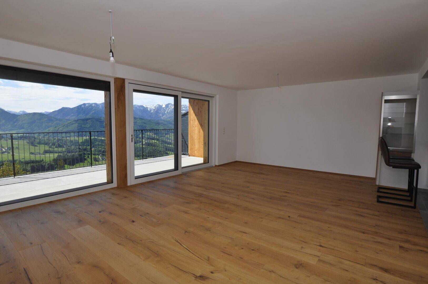 Geräumiges Wohnzimmer mit Holzboden und weitem Blick auf die grüne Landschaft und Berge.