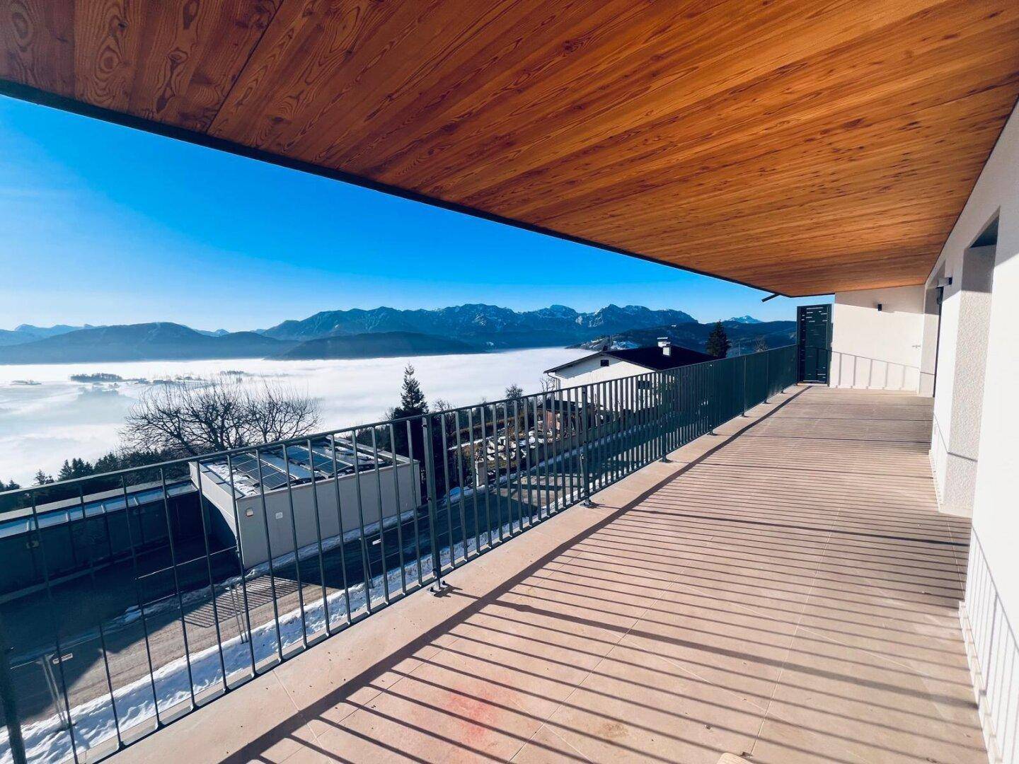 Langer Balkon mit Holzdach und weitem Blick über eine Nebellandschaft und Berge.