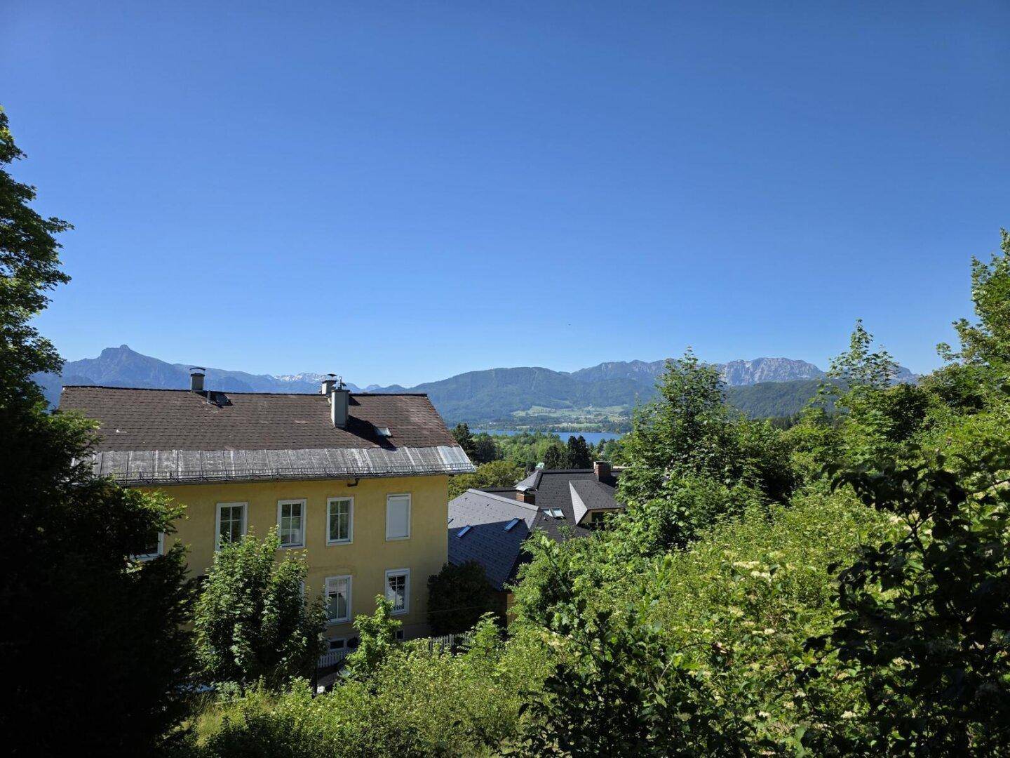 Weitläufiger Ausblick auf ein gelbes Haus, umgeben von grünen Bäumen, mit See und Bergen.