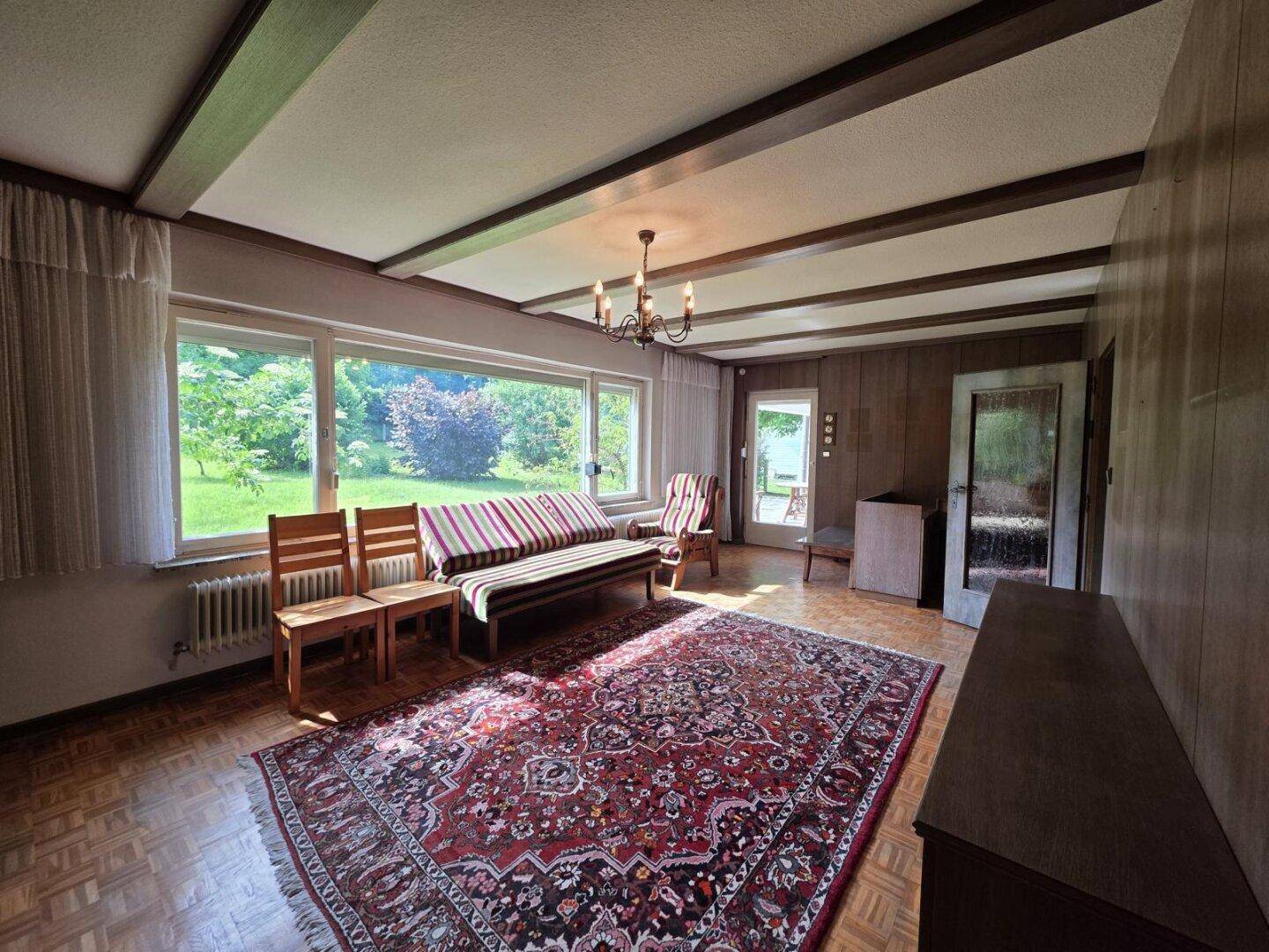 Geräumiges Wohnzimmer mit Parkettboden, Holzwänden, Deckenbalken und großem Fenster zum Garten.