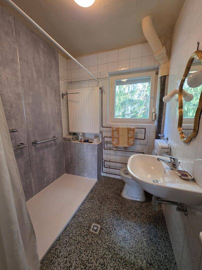 Badezimmer mit ebenerdiger Dusche, Waschbecken, WC und Fenster für Tageslicht.