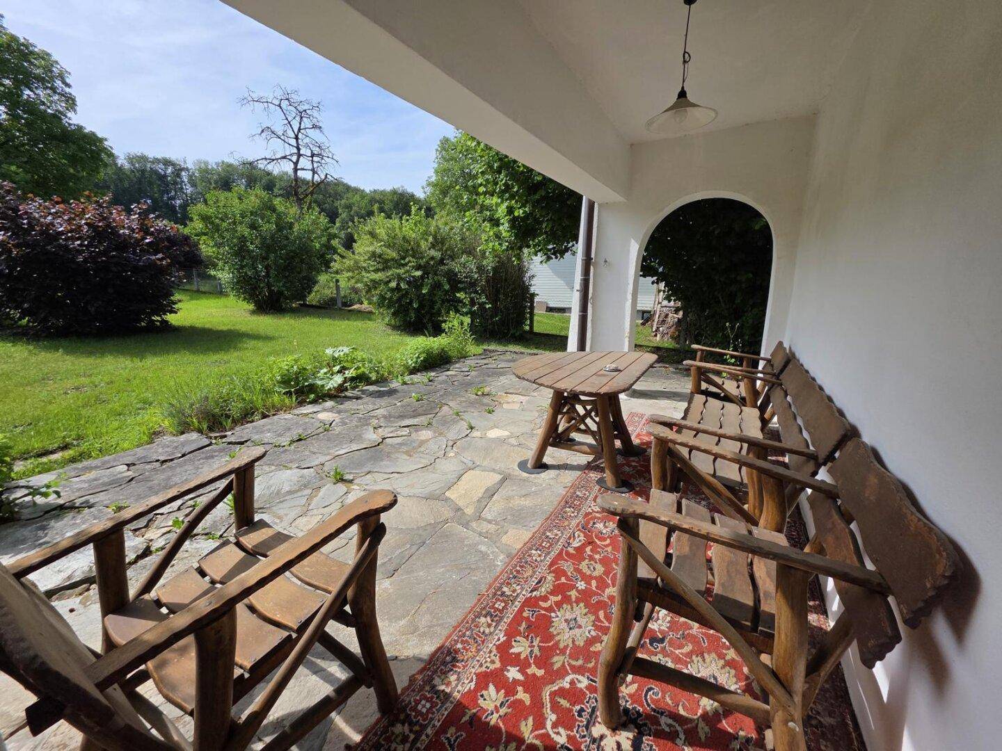 Überdachte Terrasse mit rustikalen Holzmöbeln, Steinfliesen und Blick in den grünen Garten.