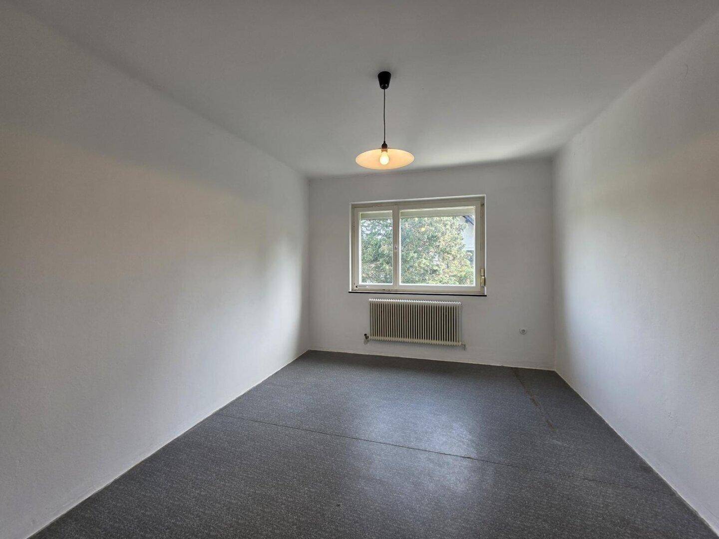 Leeres Zimmer mit weißen Wänden, grauem Boden und einem Fenster für natürliches Licht.