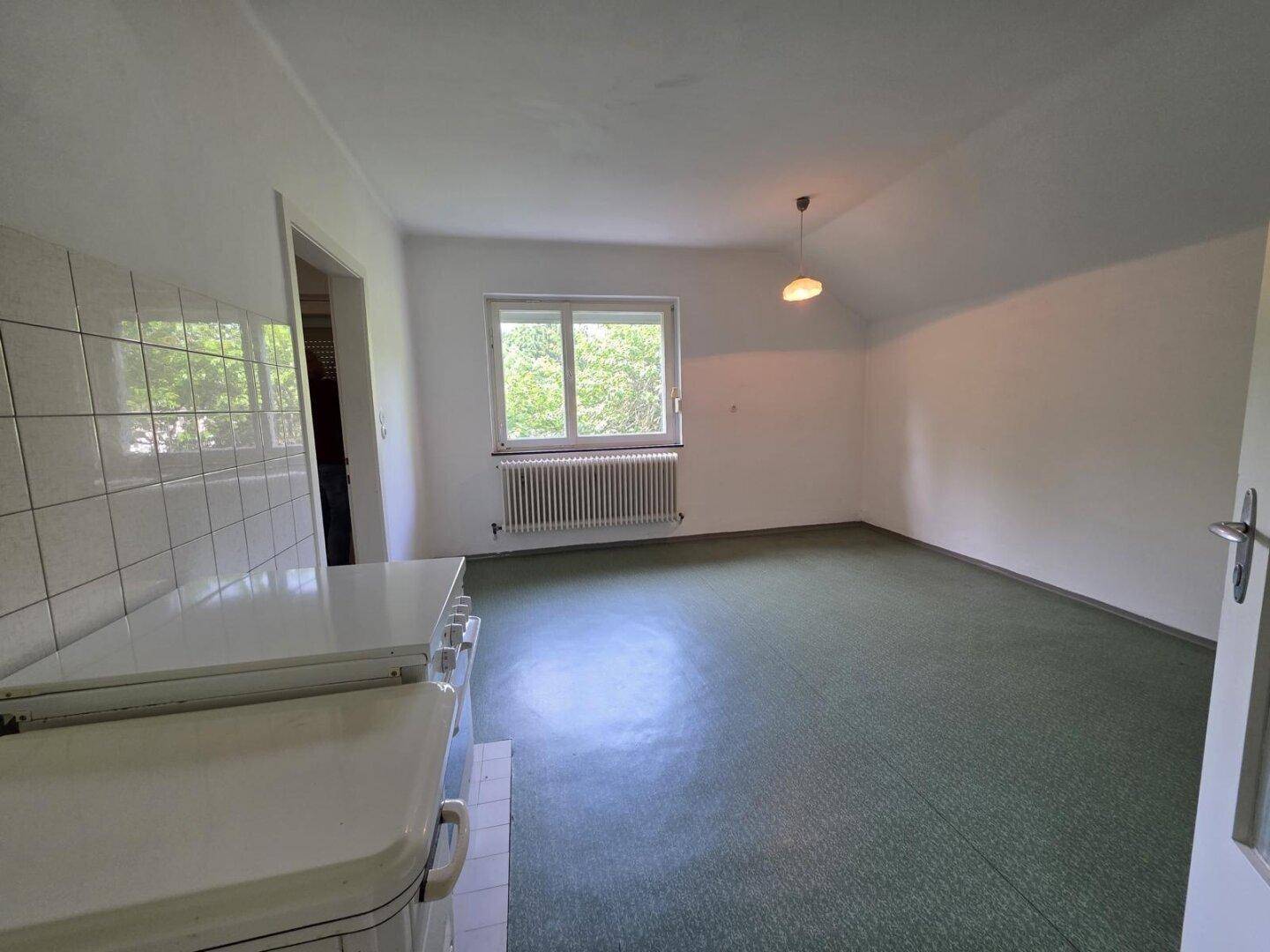 Einfaches Zimmer mit grünem Boden, Fenster und einem traditionellen Herd in der Ecke.