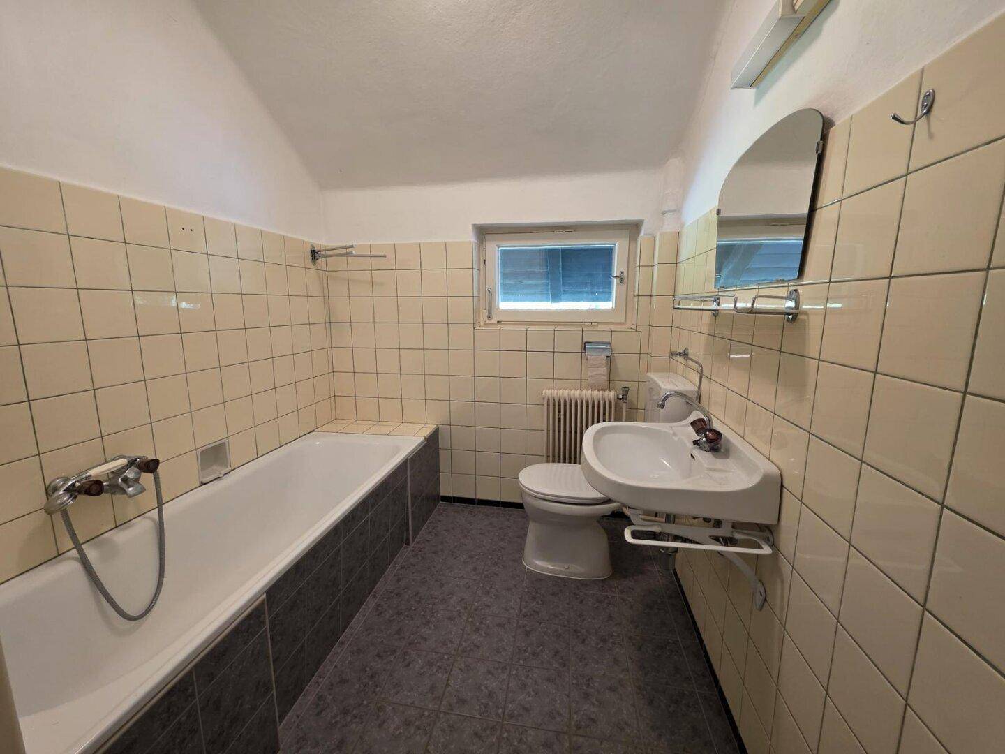 Badezimmer mit Badewanne, Waschbecken, WC und Fenster, gefliest in Beige und Grau.